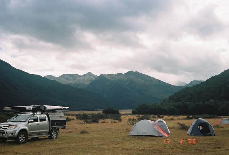 A wild backcountry camp — a kayak-topped Hilux ute and two tents pitched on a tussock flat under brooding grey mountains and a moody sky.