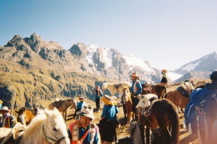 Andean horsemen and their packhorses gathered at a high pass in the Cusco region, snow-capped peaks and glaciers rising across the valley behind.