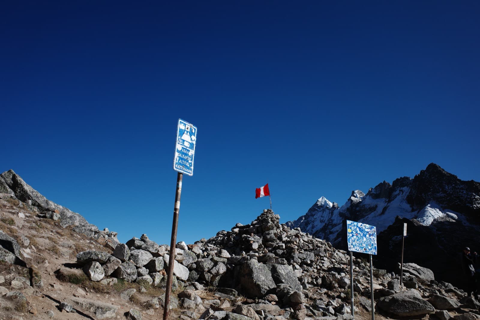 The Salkantay Pass at 4,629m — a weathered 'ABRA SALKANTAY' trail sign and a Peruvian flag on a cairn, jagged snow-capped peaks behind under a deep blue sky.