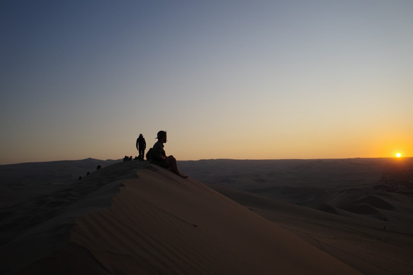 Sunset over the Huacachina dunes — travellers silhouetted on a knife-edge sand ridge as the sun drops into the desert horizon.