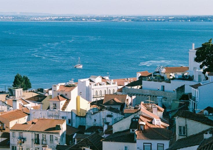 The rooftops of Alfama in Lisbon — terracotta tiles tumbling down to the Tagus, a sailboat crossing the estuary beyond.