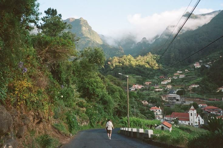 A walker heading down a quiet Madeira lane into a green mountain valley of terraced smallholdings, cloud spilling over the peaks.