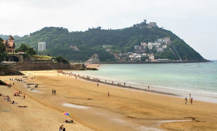 La Concha beach in San Sebastián — the wide golden crescent curving toward Monte Igueldo, calm Basque water, swimmers dotting the shallows.
