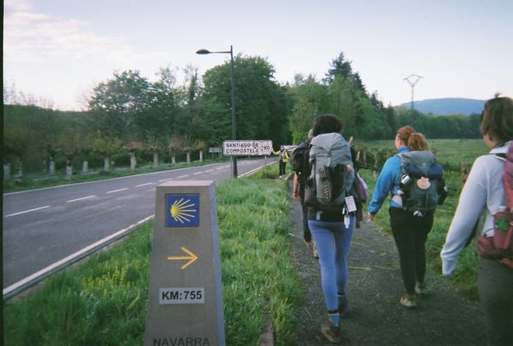 Pilgrims walking the Camino de Santiago at dawn — backpacks on, the yellow scallop-shell waymark and a road sign reading Santiago de Compostela, green Navarra hills ahead.