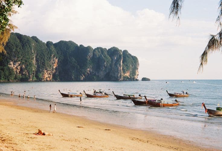 Ao Nang beach near Krabi at golden hour — a row of longtail boats moored on the sand, towering limestone karsts curving out into the Andaman Sea, swimmers in the shallows.