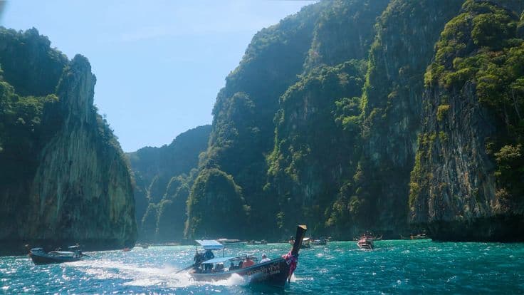 A longtail boat speeding through the turquoise water between the sheer green limestone cliffs of the Phi Phi islands, other boats dotted across the bay.