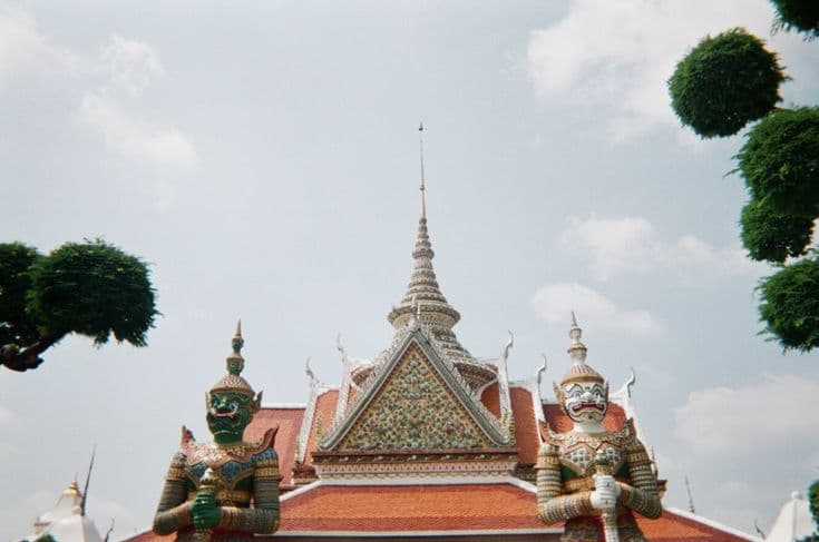 The spire of Wat Arun in Bangkok flanked by two mosaic-covered guardian giants (yaksha), the orange-tiled roofs and manicured topiary framing a bright Thai sky.