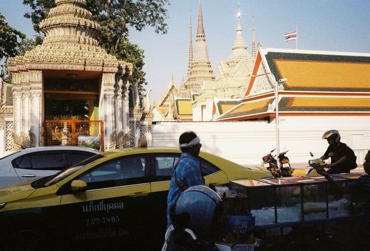 A Bangkok street outside Wat Pho — a green-and-yellow taxi, a street-food cart, a vendor in a headband and a motorbike rider, the temple's golden spires rising behind the white wall.