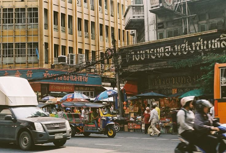 Bangkok's Chinatown (Yaowarat) by day — a tuk-tuk and motorbikes on the road, market umbrellas and old gold-shop signage in Thai and Chinese, the everyday chaos of the district.