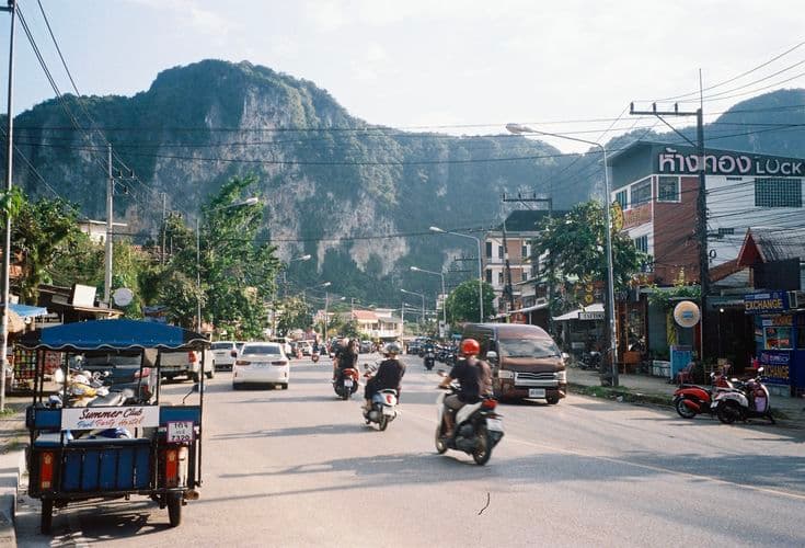 The main street of Ao Nang in Krabi — a party tuk-tuk, scooters and songthaews on the road, a dramatic limestone karst mountain rising straight up behind the shops and exchange booths.